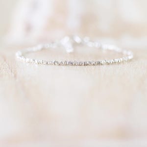 May include: A delicate silver bracelet composed of numerous small, textured, metallic segments linked together in a continuous circular chain. The surface of each individual link features a faceted, reflective finish that catches light.