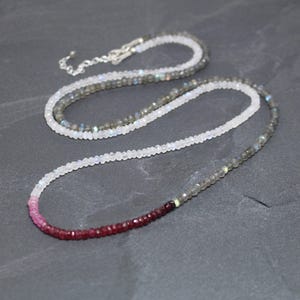 May include: A long beaded necklace featuring a gradient of small, faceted gemstones. The strand transitions from deep red garnet beads to translucent white moonstone, followed by grey labradorite beads, finished with a silver-toned metal chain.