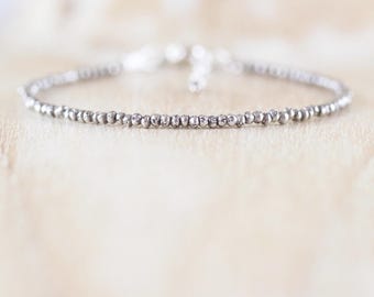Könnte beinhalten: Ein zartes Armband, bestehend aus einer einzelnen Reihe kleiner, runder, metallisch silberfarbener Perlen. Die Perlen haben eine texturierte, leicht unregelmäßige Oberfläche und das Armband verfügt über einen kleinen Metallverschluss auf der Rückseite.
