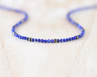 May include: A delicate necklace featuring a strand of small, faceted, deep blue lapis lazuli beads interspersed with occasional metallic, dark grey pyrite beads. The necklace is arranged in a gentle curve, showcasing the uniform size of the stones.