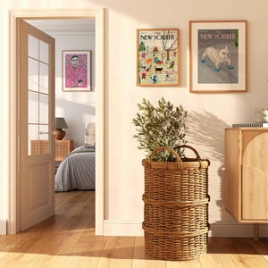May include: A well-lit interior scene featuring a large woven basket with a plant, two framed New Yorker magazine covers, and a wooden cabinet. The room has a warm, inviting atmosphere with natural light and hardwood floors.