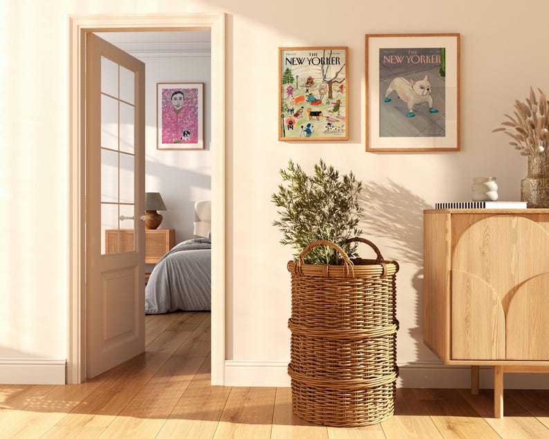 May include: A well-lit interior scene featuring a large woven basket with a plant, two framed New Yorker magazine covers, and a wooden cabinet. The room has a warm, inviting atmosphere with natural light and hardwood floors.