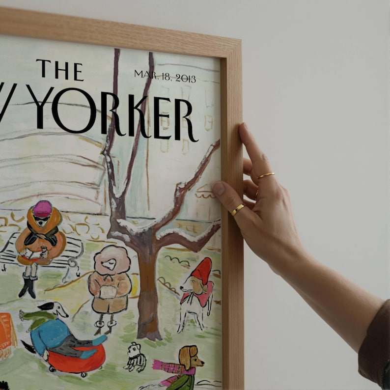 May include: A framed print of a New Yorker magazine cover from March 18, 2013. The illustration depicts dogs in winter attire in a park setting. The frame is made of light-colored wood.