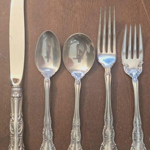 May include: A set of five silver-toned flatware pieces including one dinner knife, two spoons of varying sizes, and two forks of varying sizes. Each piece features an ornate, embossed floral pattern on the handle and a polished metallic finish.