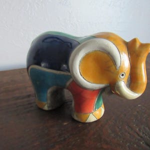 May include: A colorful ceramic elephant figurine. The elephant is painted with a patchwork design in shades of blue, green, orange, red, and white. The trunk is raised, and the ears are large and rounded. The figurine is on a wooden surface.