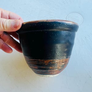 May include: A ceramic bowl with a matte black upper section and a textured, metallic copper-toned lower section featuring horizontal ridges. The rim is finished in a matching copper tone, and the base of the bowl is a speckled off-white color.