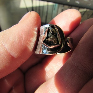 May include: A silver ring with a large, irregularly shaped, dark gray stone set in the center. The ring is designed with a twisted band.