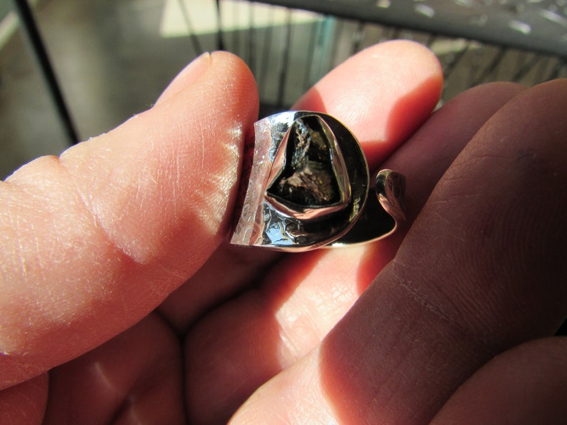 May include: A silver ring with a large, irregularly shaped, dark gray stone set in the center. The ring is designed with a twisted band.