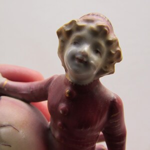 May include: A vintage porcelain figurine of a child emerging from a cracked eggshell. The child wears a burgundy outfit with gold accents. The figurine has a detailed face and is painted in soft, muted colors.