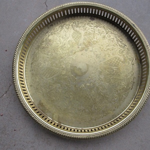 May include: A round brass tray featuring an intricate engraved floral pattern across the center surface. The tray has a raised gallery edge with a vertical slotted design and a twisted rope-style border along the outer rim of the metal piece.