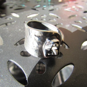 May include: A silver ring with a textured band and a dark gray, three-dimensional design. The ring is on a gray surface with a patterned design.