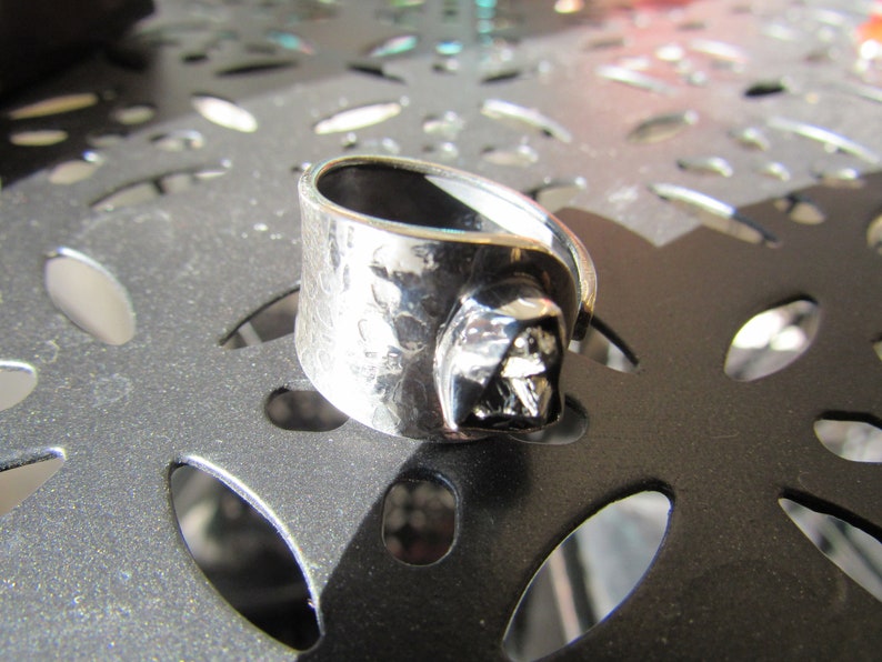 May include: A silver ring with a textured band and a dark gray, three-dimensional design. The ring is on a gray surface with a patterned design.