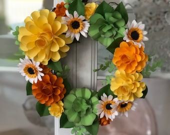 May include: A paper flower wreath with yellow, orange, and white flowers, and green leaves. The wreath is hanging on a white door.