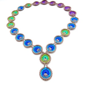 Pode incluir: Um colar de crochê colorido com um pingente. O colar é feito de fio marrom com detalhes azuis, verdes e roxos. O pingente é um círculo grande e redondo com um design azul e verde.