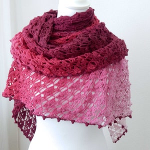 Peut inclure: Un châle en crochet dans des tons de rose et de rouge, avec un motif de dentelle délicat. Le châle est drapé sur un mannequin blanc.