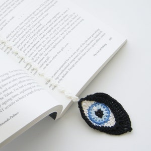 May include: A crocheted bookmark with a blue evil eye design. The eye is surrounded by white and black yarn. The bookmark is hanging from a book.