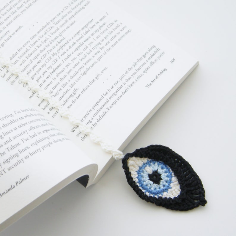 May include: A crocheted bookmark with a blue evil eye design. The eye is surrounded by white and black yarn. The bookmark is hanging from a book.