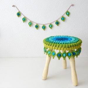 May include: A crocheted stool with a colorful, peacock-inspired design. The stool has a round, multi-colored top with a green and blue fringe. The stool is made of wood and has three legs.