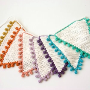 Puede incluir: Una guirnalda de crochet blanco con un arcoíris de colores. La guirnalda está hecha de triángulos con pompones alrededor de los bordes. Los colores son amarillo mostaza, naranja, rosa, morado, verde azulado y turquesa.