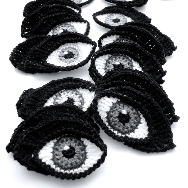 May include: A collection of crocheted black eye patches with white and gray irises. The eye patches are arranged in a staggered pattern, creating a visually appealing and unique design.