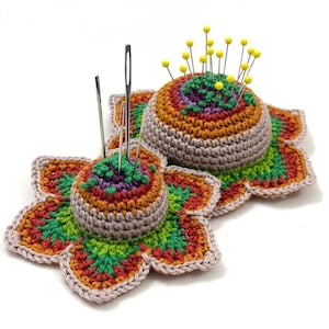 Può includere: Due cuscini per spilli fatti a mano all'uncinetto a forma di fiori. I cuscini per spilli sono al centro del fiore e sono riempiti di aghi e spilli. I fiori sono fatti di filato colorato nelle tonalità dell'arancione, verde, viola e marrone.