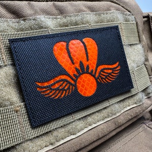 May include: A black patch with a bright orange reflective design of a winged parachute. The patch is made of a textured fabric and has a hook and loop backing.