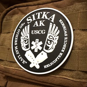 Può includere: Un cerotto rotondo bianco e nero con il testo "SITKA AK USCG HELICOPTER RESCUE SWIMMER OTHERS MAY LIVE?" in lettere bianche. Il cerotto presenta un'immagine stilizzata di due ali con un simbolo medico al centro.