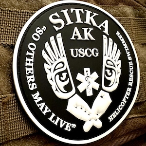 Puede incluir: Un parche redondo en blanco y negro con el texto "SITKA AK USCG" y "SO OTHERS MAY LIVE". El parche presenta un símbolo médico y alas estilizadas, con "HELICOPTER RESCUE SWIMMER" alrededor del borde. El parche está sobre una superficie marrón texturizada.