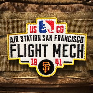Puede incluir: Un parche rectangular con el texto "AIR STATION SAN FRANCISCO FLIGHT MECH" en letras negras. El parche incluye el texto "US CG" y los años "19" y "41". Un logotipo de béisbol está en la parte superior.