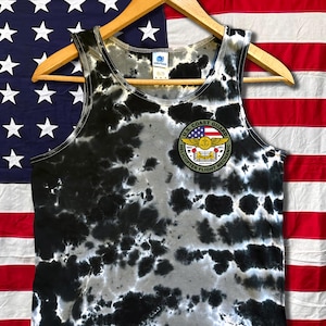 May include: A black, white, and gray tie-dye tank top with a U.S. Coast Guard Helicopter Flight Mechanic patch. The tank top is hanging on a wooden hanger in front of an American flag.