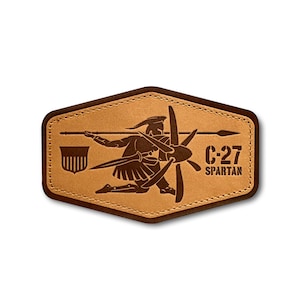 Peut inclure: Un écusson en cuir marron avec une bordure cousue. L'écusson représente la silhouette d'un guerrier spartiate tenant une lance et une hélice. Le texte "C-27 SPARTAN" est imprimé sous l'image.