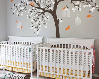 May include: A white and brown tree wall decal with white birdcages and orange birds. The decal is displayed on a gray wall above two white cribs with yellow and gray bedding.