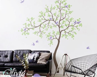 May include: A wall decal featuring a tree with green leaves and purple birds. The tree is brown and has a thick trunk. The decal is shown on a white wall in a living room setting with a black leather couch and a black wire chair.