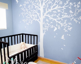 May include: A large white wall decal of a tree with spreading branches and leaves, featuring several birds perched in the branches and flying nearby, applied to a light blue wall behind a black wooden crib with a white bumper and a plush toy.