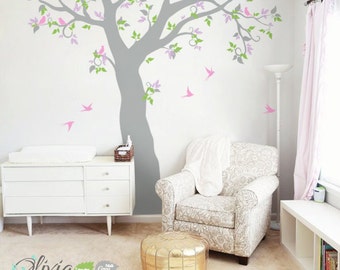 May include: A nursery room with a white dresser, a gray armchair, a gold ottoman, and a large gray tree decal with pink and green leaves and pink birds on a white wall.