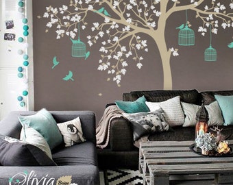 May include: A living room with a brown wall featuring a large beige tree decal with white flowers and teal birdcages. The tree has teal birds perched on the branches. The room has a gray couch with teal pillows and a wooden coffee table.