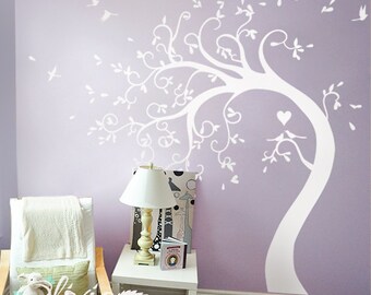 May include: White wall decal of a tree with a heart and birds on a branch. The tree has a whimsical, curly design. The decal is on a lavender wall in a room with a white chair and a lamp on a white side table.