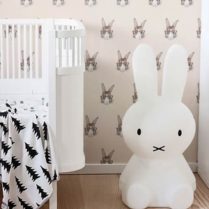 May include: A white bunny nightlight with black eyes and a black mouth sits on a wooden floor in front of a pink wall with a repeating pattern of brown rabbits wearing glasses and pink bows.