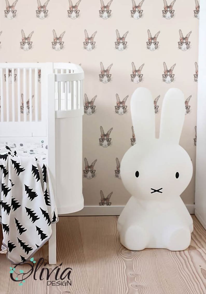 May include: A white bunny nightlight with black eyes and a black mouth sits on a wooden floor in front of a pink wall with a repeating pattern of brown rabbits wearing glasses and pink bows.