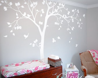 May include: A white tree wall decal with birds and leaves, a changing table with a pink and white patterned pad, a white wicker basket with a white towel, a small wooden dresser with three drawers, a small wooden shelf with a picture frame and a pink and white storage bin, and a rocking chair with a pink and white throw blanket.