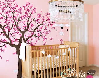 May include: A pink and brown nursery with a crib, a chandelier, and a wall decal of a tree with pink leaves. The text "Olivia Design" is on the floor in front of the crib.