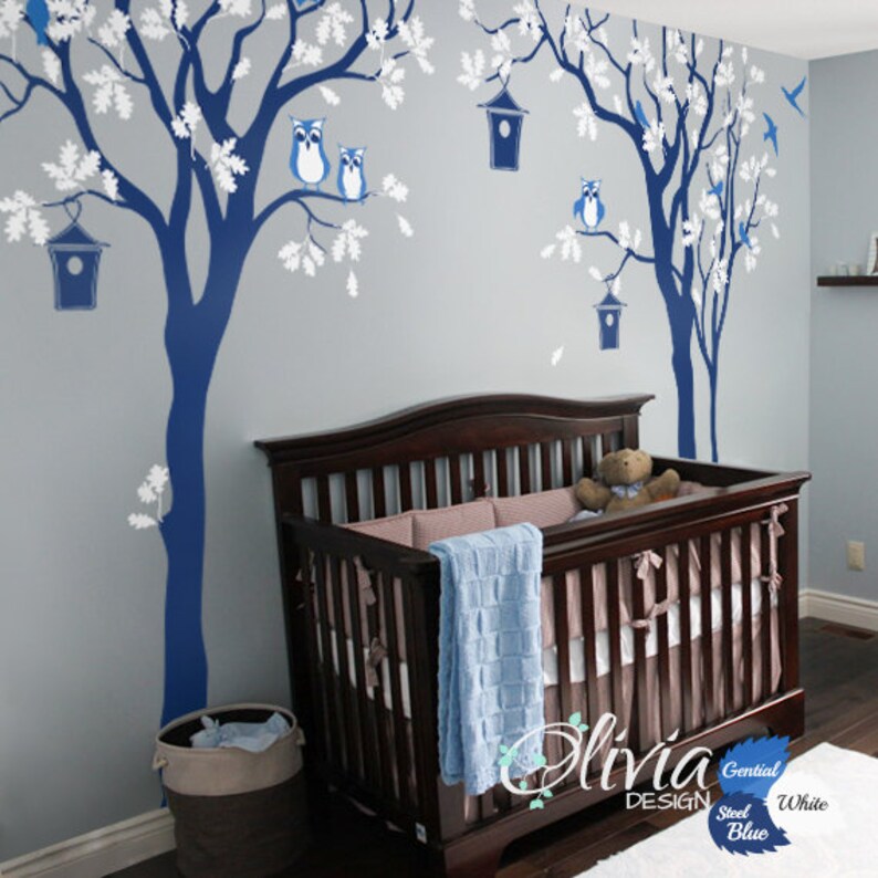 Pode incluir: Um quarto de beb&eacute; com um ber&ccedil;o e um mural de parede com duas &aacute;rvores azuis com ramos brancos, p&aacute;ssaros azuis e corujas azuis. As &aacute;rvores t&ecirc;m casas para p&aacute;ssaros penduradas nos ramos. O ber&ccedil;o &eacute; de madeira castanho e tem um cobertor azul e um urso de peluche dentro.