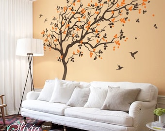 May include: A white couch with pillows in a living room with a large wall decal of a brown tree with orange leaves and silhouettes of birds flying around it. The text "Olivia Design" and "Brown Rust Orange" is visible in the bottom left corner of the image.