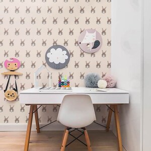May include: A white desk with a white chair and a light pink rug. The wall is covered in a wallpaper with a repeating pattern of brown bunny faces wearing glasses. There are two round wall decorations, one with a cloud and the other with a fox.