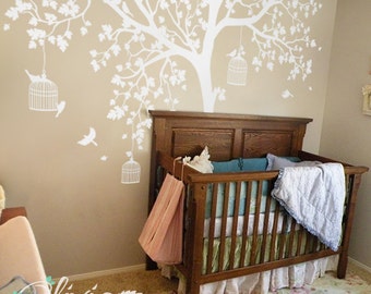 May include: White tree wall decal with birds and hanging birdcages above a wooden crib with a white and blue blanket and a pink and white bag hanging on the side.