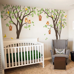 Pode incluir: Um ber&ccedil;o branco com um colch&atilde;o verde em um quarto de beb&ecirc; com uma poltrona cinza e um pufe marrom. A parede &eacute; decorada com um adesivo de &aacute;rvore com corujas e casas de p&aacute;ssaros.