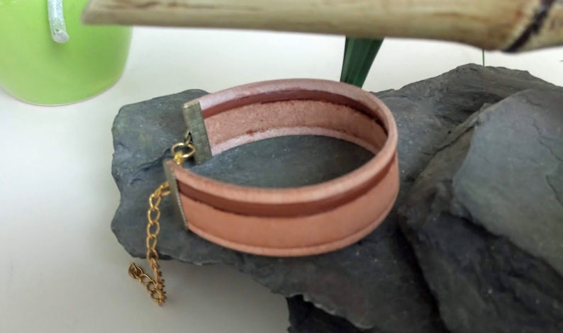 May include: A light brown leather bracelet with a gold chain and clasp. The bracelet has a brown and white striped design.