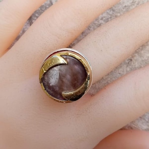 Può includere: Un anello rotondo con una grande pietra ovale viola chiaro. La pietra è incastonata in una montatura in metallo color oro con accenti curvi. La fascia dell'anello è color argento.