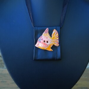 Pu&ograve; includere: Una collana con un ciondolo a forma di pesce in ceramica, smaltato in arancione, rosa e giallo. Il pesce ha due bottoni neri per gli occhi ed &egrave; attaccato a un nastro di raso nero.