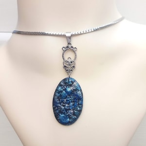 May include: A silver-toned necklace with an oval, blue pendant. The pendant has a textured, mosaic-like appearance. The necklace is displayed on a mannequin bust, showcasing the jewelry's design and details.